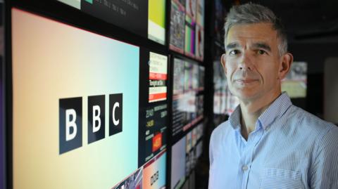 Matt Brittin standing in front of a bank of screens, including one with the BBC logo