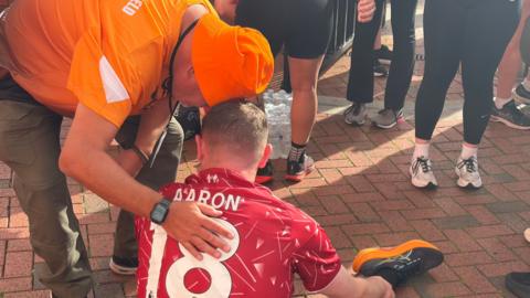 Shaun McEntee sits on the floor wearing a red Liverpool FC shirt with Aaron 18 on his back. A man in an orange t-shirt and orange bucket hat rests his head on top of Shaun's. 