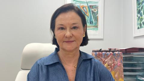 A woman with dark bobbed brown hair, wearing a blue open collared shirt and a gold necklace and gold stud earrings. She is sitting on a white leather office chair and there are a stack of well-ordered folders behind her, as well as a scientific model of the skin. Behind her are two abstract painting on a white wall.