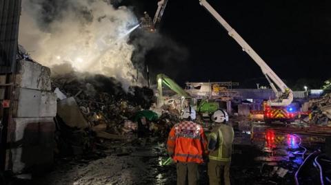 An aerial firefighting ladder and firefighter can be seen above a pile of smoking waste. Other fire crews are in the foreground