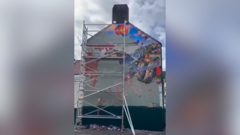 The large wall of a building is pictured with parts of a colourful Mural removed. In front of the wall is scaffolding.