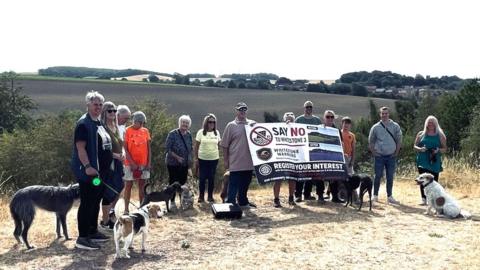 A group of people and dogs are standing surrounded by fields. They are holding a large banner opposing a solar farm