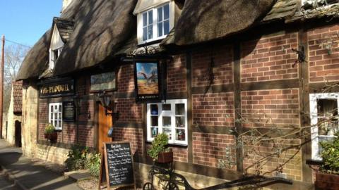 A traditional pub built out of bricks with a thatched roof. There is a sign with a plough painted on it on the front, and a sandwich board with chalk writing on it.