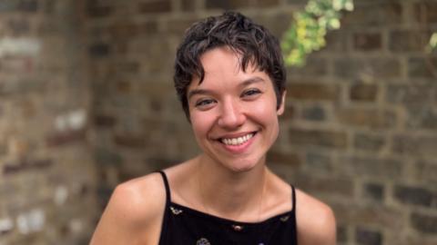 The image shows a medium close-up portrait of a woman short, dark brown hair.  She is smiling broadly, showing her teeth, and looking directly toward the camera. She is wearing a black top with small, scattered embroidered patterns.  