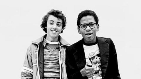 A black-and-white photo of two boys who are smiling at the camera in 1979. The boy on the left is wearing a striped shirt and a jacket, while the boy on the right is wearing a white tshirt with a cartoon cowboy, a black jacket and he is wearing black glasses