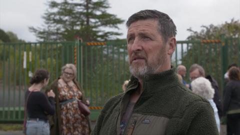Michael Phillips, a middle aged man with short brown hair and a beard, wearing a green fleece. A group of people can be seen behind him with a large metal gate in the background