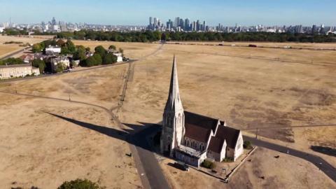 Aerial view of a church surrounded by drought-hit grassland, with central London visible in the distance