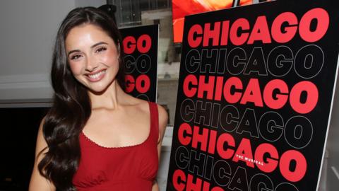 Reece wears a bright red dress and has long brunette hair which sits past her shoulders. She stands in front of a Chicago The Musical sign