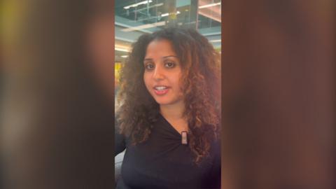 A young woman with long curly brown hair talks into the camera. She is wearing a black top.