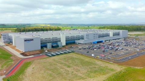 A sprawling grey box building, which is four storeys high, stands next to a car park. The building has an Amazon sign in the corner and is surrounded by fields.
