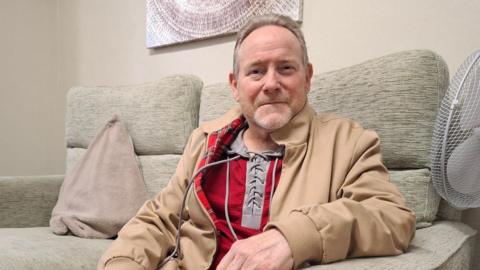 Nicholas Lawrence sits on a grey sofa while looking into the camera. He is wearing a brown overcoat and a red undershirt with grey trim.