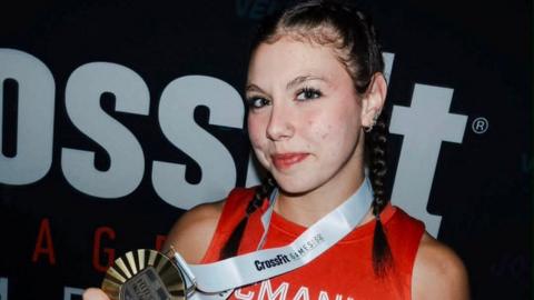 Keira McManus, a girl with dark hair tied in two plaits and wearing a red sports top, smiles as she holds up a large gold medal, which is around her neck.