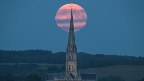 A pink-coloured full Harvest Moon rises behind Salisbury Cathedral spire. There are hills and trees in the distance and streaks of grey cloud across the Moon