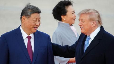 President Xi smiles and looks toward Trump, who has his hand on Xi's shoulder. Both are in suits and their haor is slightly tussled by wind. Xi's wife, in a light blue coat, looks away in the background. 