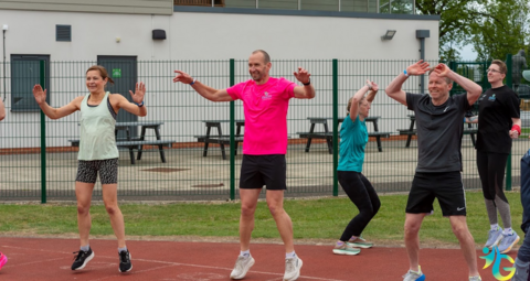 Tim is on an athletics track with a few other people, all in sports gear. He is wearing a bright pink t-shirt and black shorts. There is a patch of grass behind, and a concreted area in the background with several picnic tables in front of a sports building