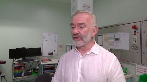 A man with grey hair and beard wearing a pink shirt stood in a hospital room
