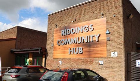 Exterior view of the Riddings Community Hub, which is a brick building with a large wooden panel mounted on the front wall displaying the text “RIDDINGS COMMUNITY HUB” in large grey letters. Two parked cars are visible in the foreground.
