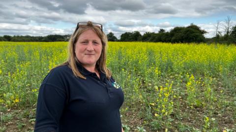 Farmer Debbie Wilkins is pictured standing in a field full of yellow flowers on a cloudy but sunny day. She has long blonde hair and a pair of sunglasses is perched on her head. She is wearing a navy blue polo shirt.