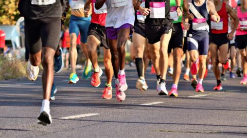 The legs of about a dozen runners wearing shorts and trainers of various colours during a running event. They are on a road and part of a bush is on the far left.