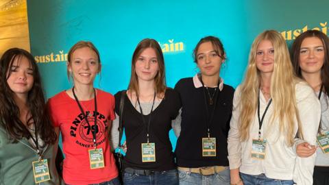 A group of teenage girls standing in a line smiling at the camera. All wear jeans and tops with lanyards for the conference.