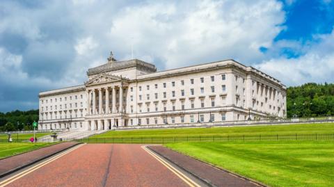 Stormont is a large cream coloured building with a number of white rectangular windows and is at the top of a hill.