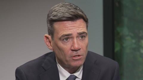 Andy Burnham has short dark hair and wears a dark suit. He is mid-speech and looks slightly off to the side