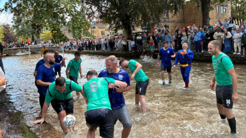 About a dozen men are playing football in a shallow, wide river in the middle of a town. They are wearing turquoise or blue shirts and the referee is in a superman outfit. One man is tackling another player in the foreground and the football is in mid-air. There are lots of spectators on the river bank