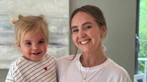 Emily is carrying Leni, who is blond. Both are wearing white t-shirts with Leni's striped with a heart.