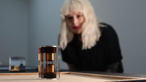 A blonde woman dressed in black leans forward to look at a sculpture by artist Tanoa Sasraku. The sculpture is made from found objects including brass paperweights and a glass casing.