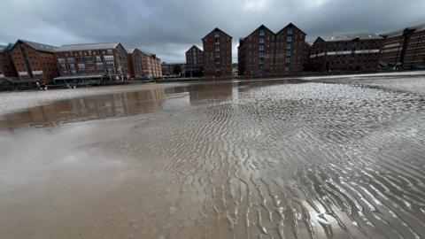 A photo showing the canal basin at Gloucester Docks. It shows ripples of mud breaking over the surface of the water. It stretches out quite far, but to the left the silt gives way to deeper water. On the other side of the dock you can see the traditional red brick warehouses under a cloudy sky.