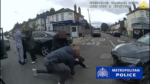 Police bodycam footage shows two officers arresting a man, who is on his hands and knees on the road, beside some parked cars in a residential street. To their left, are three other individuals, their faces blurred. 