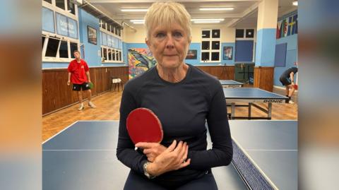 A woman with grey hair and wearing a black T shirt, holding a red table tennis bat while sitting on the edge of a table tennis table 