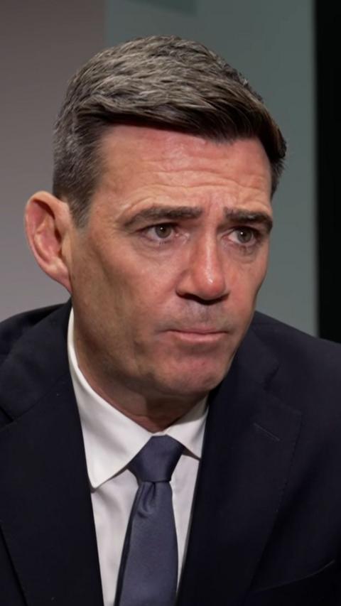Andy Burnham has short dark hair and wears a dark suit. He is mid-speech and looks slightly off to the side