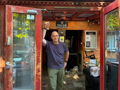 A man stands in front of two open doors that lead to a reception area. He is leaning with one arm against one of the doors. There is a sign behind his head that reads "Physical Culture". He is wearing a dark blue T-shirt and khaki green trousers. In the background there are two black sofas and a fan on the floor, along with other furniture. 