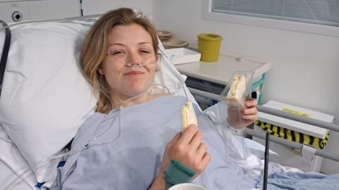 A woman in a hospital wearing a surgical gown. She has tubes up her nose and is holding a cheese sandwich in a packet, smiling