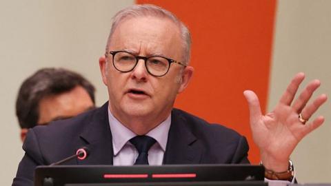 Anthony Albanese, wearing a suit and black glasses, speaks while holding his hand up, against a beige and orange background