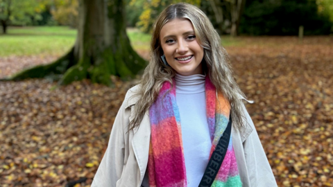 A young woman with long blonde hair, a multi-coloured scarf, a turtle neck white top and a beige coat. She's standing in front of a tree, with lots of autumnal leaves on the ground. 