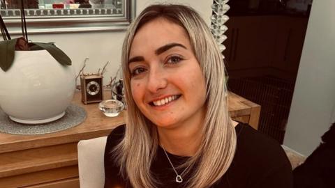 Maddy Cusack pictured smiling at home. She has long blonde hair worn loose and wears a silver heart necklace over a black top. She's pictured inside, in front of a side board with ornaments and plants 