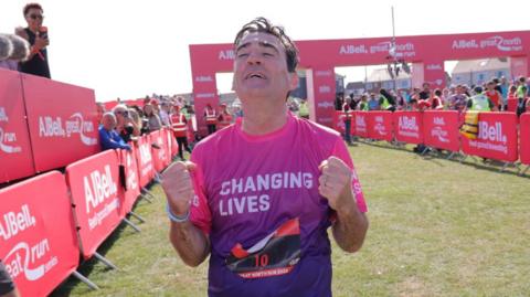 Andy Burnham crossing the finishing line in South Shields. He is clenching his fists and looking upwards with an expression of strain, and wearing a pink and purple T-shirt with changing lives on it. Behind him stretch out the hoardings with crowds of spectators, and another runner leans on it to the left, with two more standing on the right.