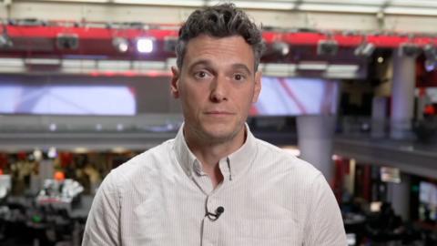 A man wearing white and beige striped shirt, with a clip-on mic. He has short dark grey hair.