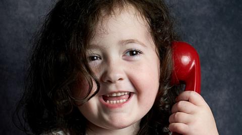Evie is smiling and holding a red phone. She is a young girl with long thick dark hair