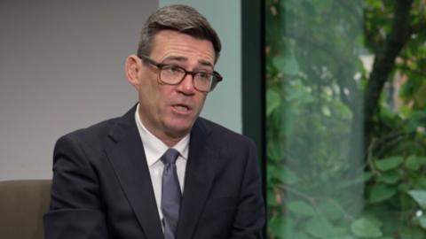 Andy Burnham, who has short dark hair and black glasses, wears a white shirt and a navy blazer