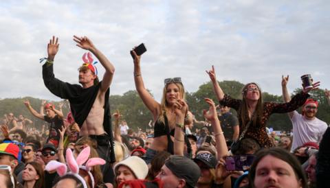 Crowds of festival-goers wave their hands in the air at Leeds Festival. There a few people in the crowd raised up by sitting on the shoulders of other people. 