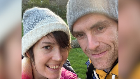 The image shows a woman and a man taking a selfie outdoors, both wearing grey woolly hats. The woman, on the left, has a brown fringe and is wearing a pink coat, while the man, on the right, is dressed in a yellow hoodie and has light stubble. They are standing close together and smiling for the photograph.