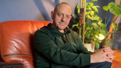 A man with a bald head in a green zip-up top sits with his fingers interlocked on his knee on a brown chair with a rubber plant in the background