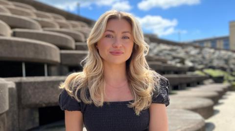 Daisy is sitting on curved concrete seating steps outdoors in a coastal setting, facing the camera. She's wearing a dark short-sleeved dress and a wristwatch. Behind her are layered, rounded concrete structures leading toward a rocky shoreline. The sky is bright blue with scattered white clouds, and sunlight casts soft shadows across the textured concrete surfaces.