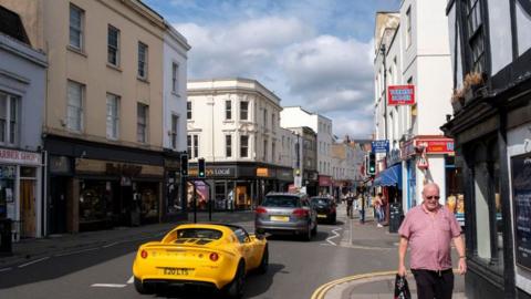 A high street with cars driving down the road with shops, food outlets and on either side. A man wearing a red shirt and sunglasses is walking on the side of the street. 