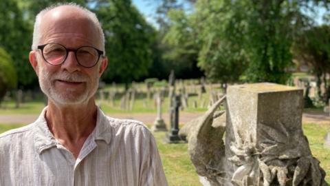 Adrian Bean, standing in a graveyard, by stones, wearing a cream top, glasses and he has short grey hair and a beard.