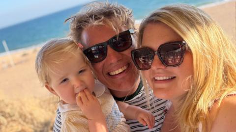 Lily is on a beach with her dad, Jonny, and her mum, Rosie.  The beach and sea are in the background.  Lily is in a sundress; Jonny and Rosie are wearing sunglasses and are smiling at the camera. 