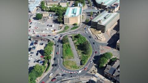 An aerial view of a roundabout with cars driving round it. The roofs of buildings and a car park dot the landscape along with a few areas of trees and grass.